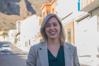 Mujer sonriente con chaqueta, de pie en una calle con casas blancas y montañas al fondo.